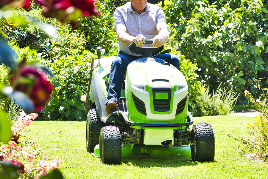 Senior man 75 old years driving a tractor lawn mower in garden with flowers. Green and white ride on mower, turning in field between colorful flowers