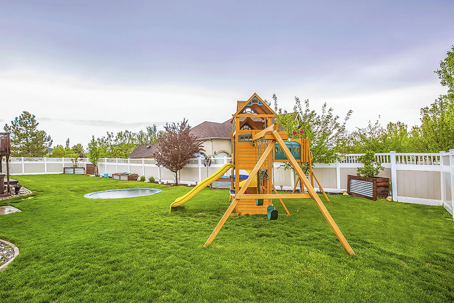 Playground equipment and small pool at the spacious backyard of a house. Houses and trees against cloudy sky can be seen in the background.